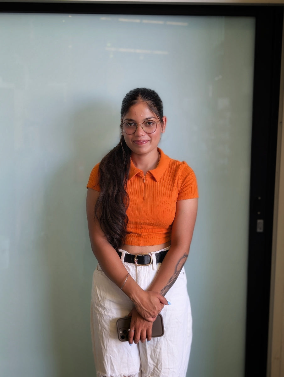 Woman wearing an orange shirt and white pants standing against a light-coloured wall.