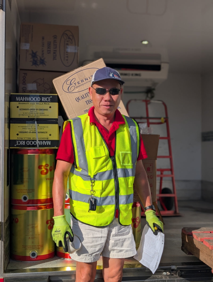 Man in a warehouse wearing a safety vest and gloves, standing in front of stacked boxes and a ladder.