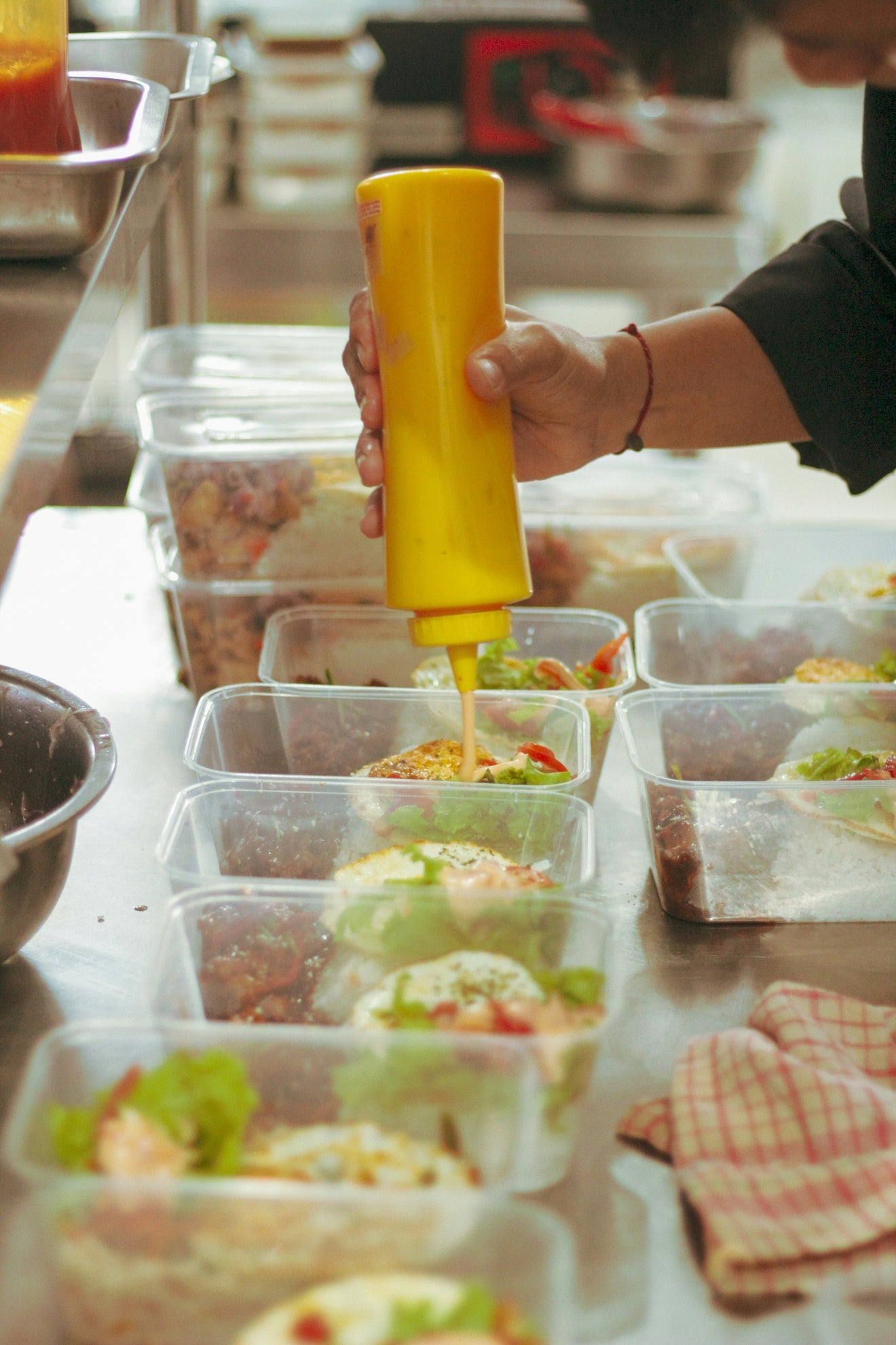 Person pouring yellow sauce into prepped food containers in a kitchen setting